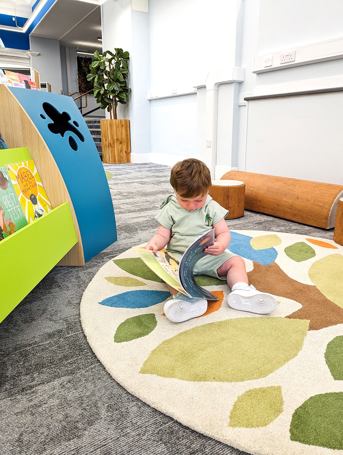 toddler reading picturebook on tree rug with library furniture