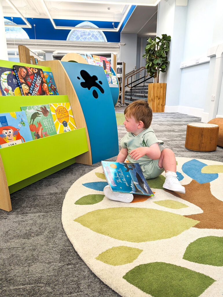 toddler sitting on a tree rug and looking at books in children's library bookcase